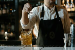A bartender in a white shirt and apron stirs a whiskey cocktail on a bar counter.